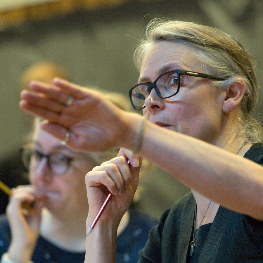 19-WhenWeHaveSufficientlyTorturedEachOther-KatieMitchellRehearsal-Stephen-Cummiskey-2000x1000 An image of Katie Mitchell in rehearsal, holding a pencil in her right hand and gesturing with her left hand towards something happening in front of her.