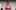 2024_New Views_Here to Help_Rehearsed Reading_Emma Hare A person with long brown braided hair, dressed in a pink hoodie, stands holding a few sheets of paper. The background is plain, mostly grey, with some partially visible elements. The person appears to be reading or presenting something.
