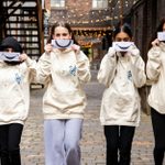 Four people stand in a cobblestone alley, wearing beige hoodies with a blue and black logo. They each hold a paper with a large, drawn smile covering their faces. String lights are visible in the background.