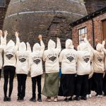 A group of people stand with their backs to the camera, all wearing matching beige hoodies with 