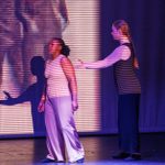 Two performers are on a dimly lit stage, with one person slightly in front of the other. Their shadows are projected on the backdrop, which is illuminated by horizontal lines of light, creating a striped pattern across their bodies and the background. Both people appear engaged.