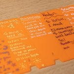 A few orange plastic planting guides on a table, labeled with various names and their care instructions written in white marker. The notes include sunlight requirements, spacing, and soil depth for plants with the names of people like Diana, Phoebus, Avistabadu, Kaden and Scurra.