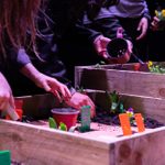 People are seen planting seedlings in wooden raised beds. They are using small garden tools and water cans. The planting area has colorful plant markers and some small potted plants. The scene appears collaborative with multiple hands working together.