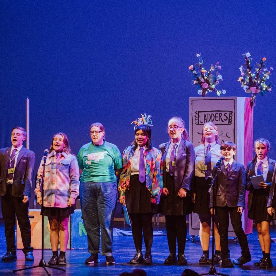 A group of people stands on stage in a theatrical performance. The performers are dressed in school uniforms and casual clothes, with some featuring colourful accessories. The background includes a blue backdrop and stage props, including a sign reading 