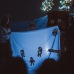 Four people are performing shadow puppetry using a backlit blue screen, displaying abstract figures. The scene is dark with illuminated tree decorations in the background, creating a whimsical, storytelling atmosphere.