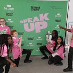 A group of six students pose enthusiastically in front of a green backdrop with the words 