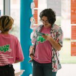 Two people standing in a room with posters on the walls and windows. One person with short hair and glasses is reading from a paper card, wearing a floral shirt over a pink t-shirt. The other person has tied-back hair and a pink t-shirt with 