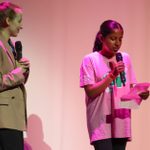 Two young women are on a stage holding microphones. The woman on the left has light hair and is dressed in a beige blazer. The woman on the right has dark hair, is in a pink Speak Up T-shirt, and is reading from a piece of paper. The background is lit with pink and purple lights.