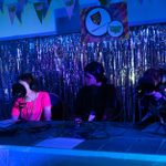 A group of five people sit at a table with microphones and headphones, looking like they are recording a podcast. The setting has blue lighting, silver streamers in the background and Speak Up bunting. The table is covered with a light cloth with equipment and notes on it.