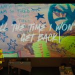 An art exhibit featuring a large screen with colorful graffiti-style artwork and text that reads, 