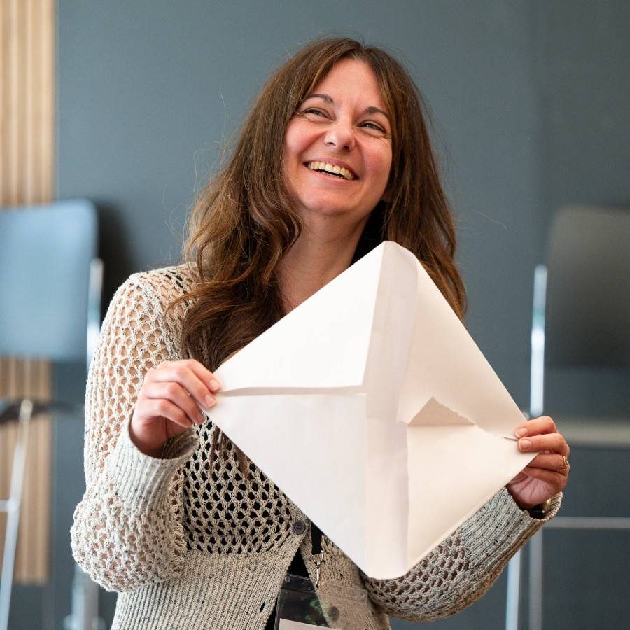 2024_Theatreworks_Influence&RapportOpenCourse_Paper_JimmyLee_2000x1000 A woman with long brown hair, wearing a crocheted beige sweater, is smiling broadly while holding up an empty white piece of paper, folded like an envelope. She is standing indoors, with grey and beige furniture in the background, resembling a rehearsal room.