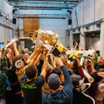 A large group of people collectively holding up a sculpture made of cardboard and paper