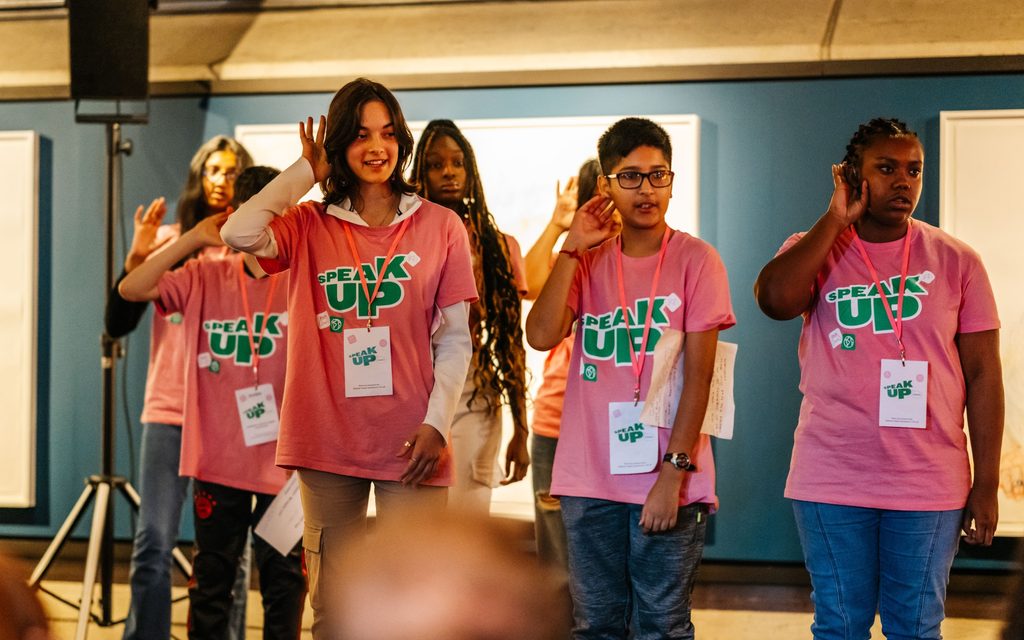 A group of young people wearing pink t-shirts with the words 'Speak Up' written on them - they are speaking British sign language