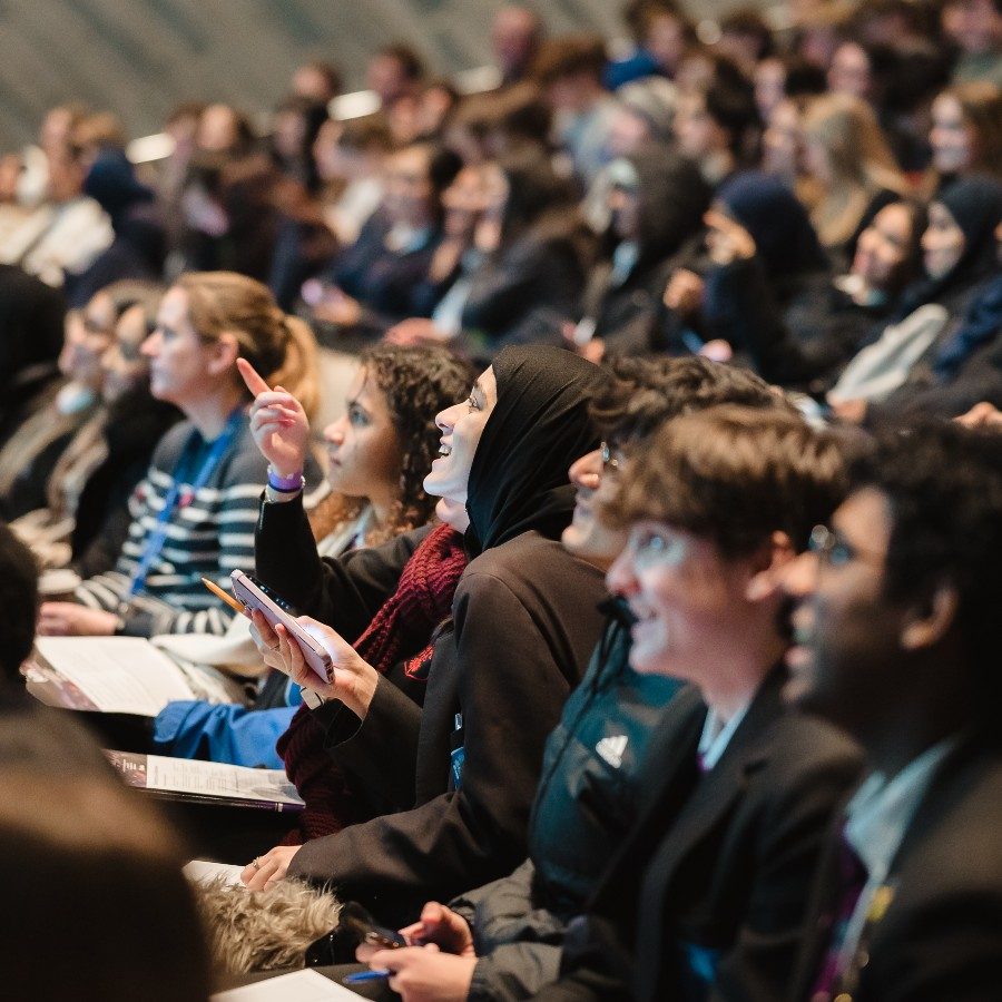A young person pointing to the stage whilst another person smils and looks upwards, they are sitting in the Lyttelton Theatre which is full of other young people