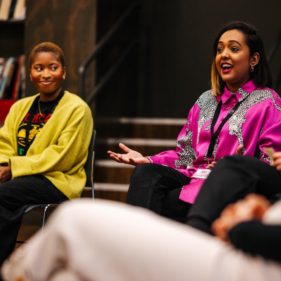 Two women sitting on chairs in a rehearsal room in a circle with other people. The woman on the right is speaking and seems to be explaining something to the group. The other woman looks at her and listens.