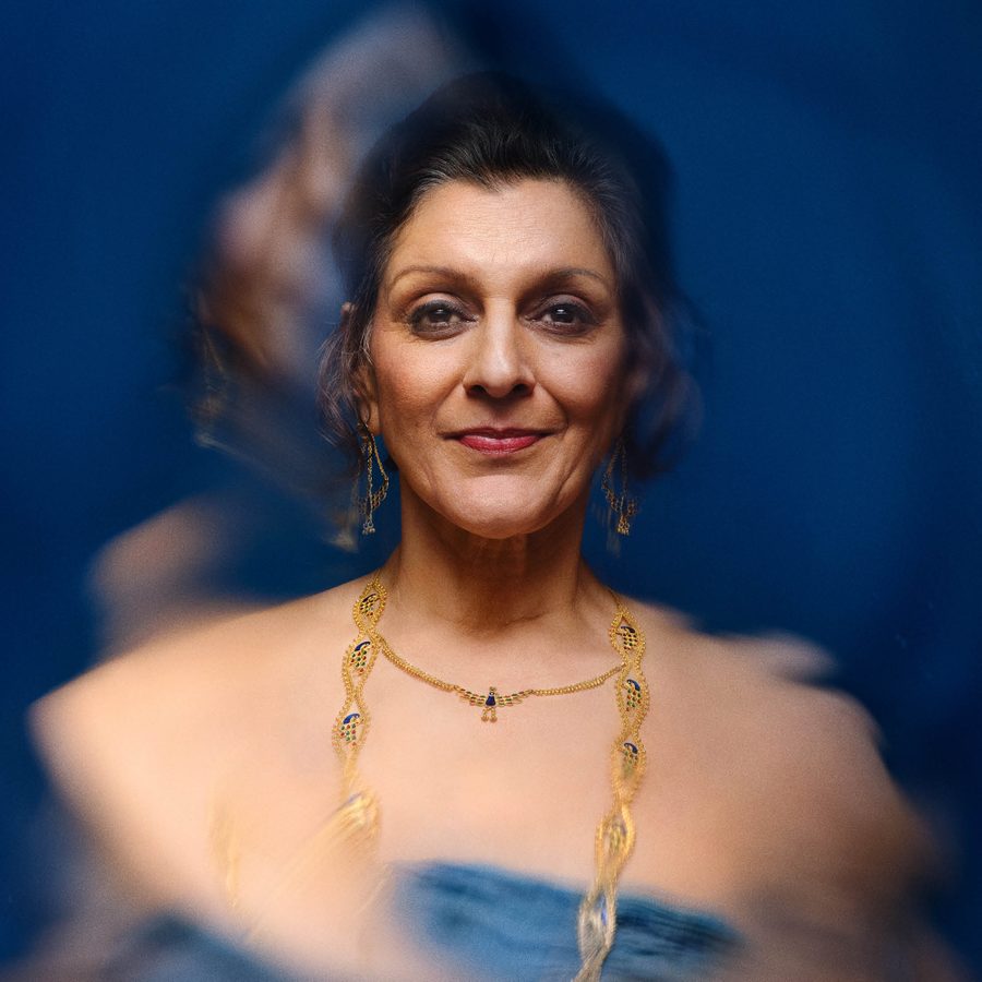 A-Tupperware-of-Ashes-National-Theatre-2000x1000 A blurred or shaken photo of a woman (Meera Syal), smiling and wearing gold jewellry and a blue dress.