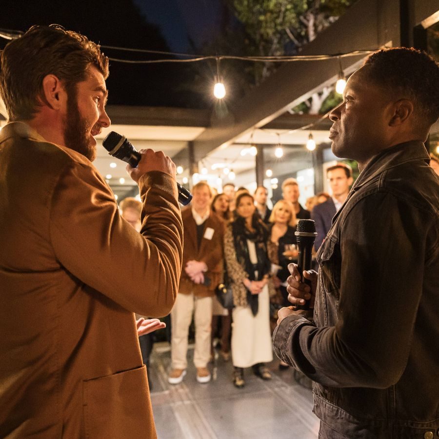 AANT-CAA-Cocktail-Party-speakers-with-Andrew-Garfied-David-Oyelowo-2000x1000 Andrew Garfield speaking into a microphone, standing next to David Oyelowo, with an audience standing in the background.