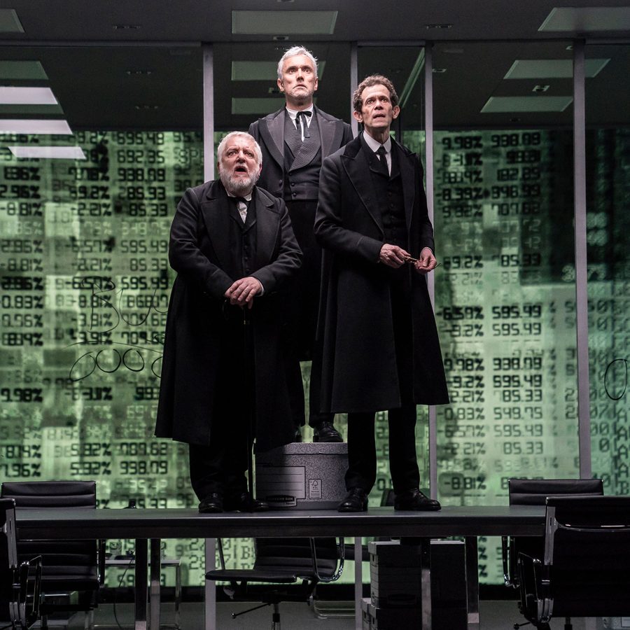 AANT-HERO-IMAGE-The-Lehman-Trilogy-2019-PRO-13-Stephanie-Berger--2000x1000 Three actors in period attire stand on stage, with a digital backdrop displaying streams of alphanumeric characters.