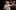 AANT-MEMBER-Hadestown-2018-PRO-19-Helen-Maybanks-2000x1250 A performer in a stylish suit smiles and gestures with one hand on his chest and the other raised. A band is visible in the dimly lit background.