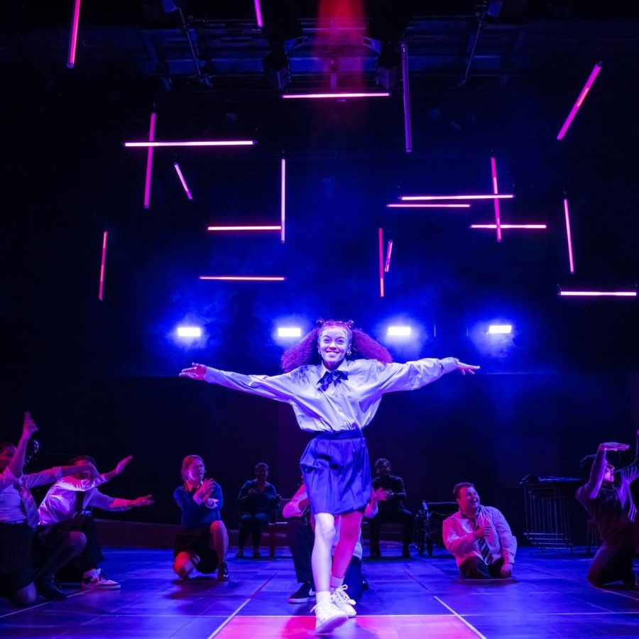 Alex-Jarrett-and-company-Our-Generation-National-Theatre-Photo-Johan-Persson_1996x998_acf_cropped Alex Jarrett steps forward, centre-stage front, arms outstretched and smiling, with members of the company sat or crouching behind her.