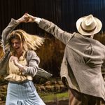 Tom Riley as Gerry spins Alison Oliver as Chris. Tom wears a suit and hat and Alison wears a blue skirt, cream blouse, and grey cardigan.