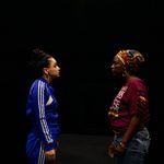 Two women, one in a colorful headdress with gold hoops, and the other in a navy Adidas tracksuit, are facing each other.