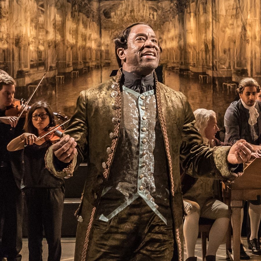 Lucian Msamati in Amadeus A man stands, smiling, hands lifted slightly. A group of people stands behind. All are wearing costumes based on the 18th century.