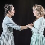 Two young women in light blue dresses facing each other. They are holding each other's hands with emotional expressions.