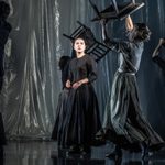 A young woman in a black dress at centre stage. Around her, four other cast members carry tables and chairs above their heads.