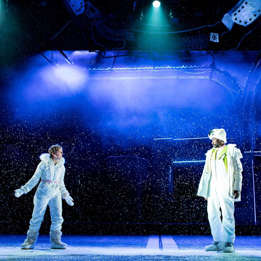 Angels-In-America-Part-1-2000x1000 Two people in white, winter attire stand on a snow-covered stage beneath blue and purple lights. They face each other, surrounded by falling snow, creating a dramatic and immersive scene.