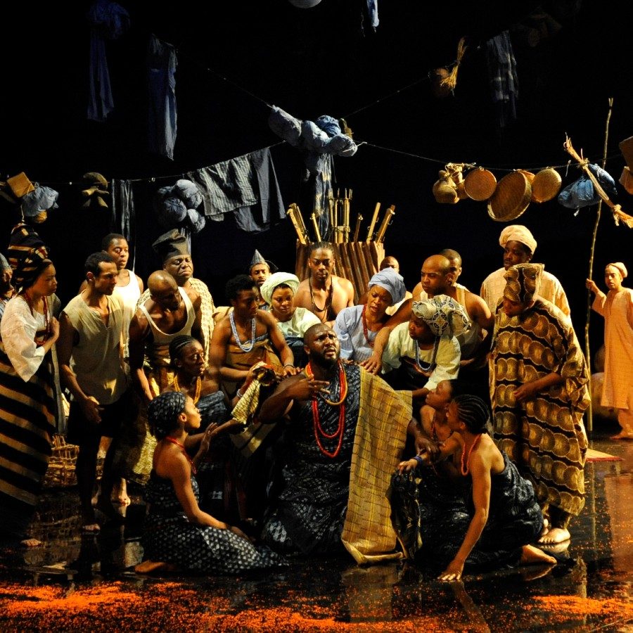 Archive_Death and the King's-2009-PRO-10-Nonso Anozie as Elesin,Giles Terera as Praise Singer_Photo Robbie Jack_2000x1000 A group of people on stage in colourful attire, set against a dark background. They are surrounded by props like baskets and cloths hanging above them. The scene conveys a sense of community or celebration.