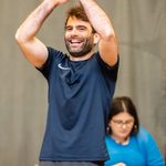 A man with brown hair wearing a navy gym shirt and grey shorts, raising his arms above his head to clap with a big smile. Behind him, a woman in a blue top is sat at a table making notes.