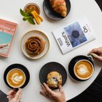 A round white table top with two cups of cappucino, three plates, each with a pastry, the book A Vision of the World by John Cheever and a What's on Guide.