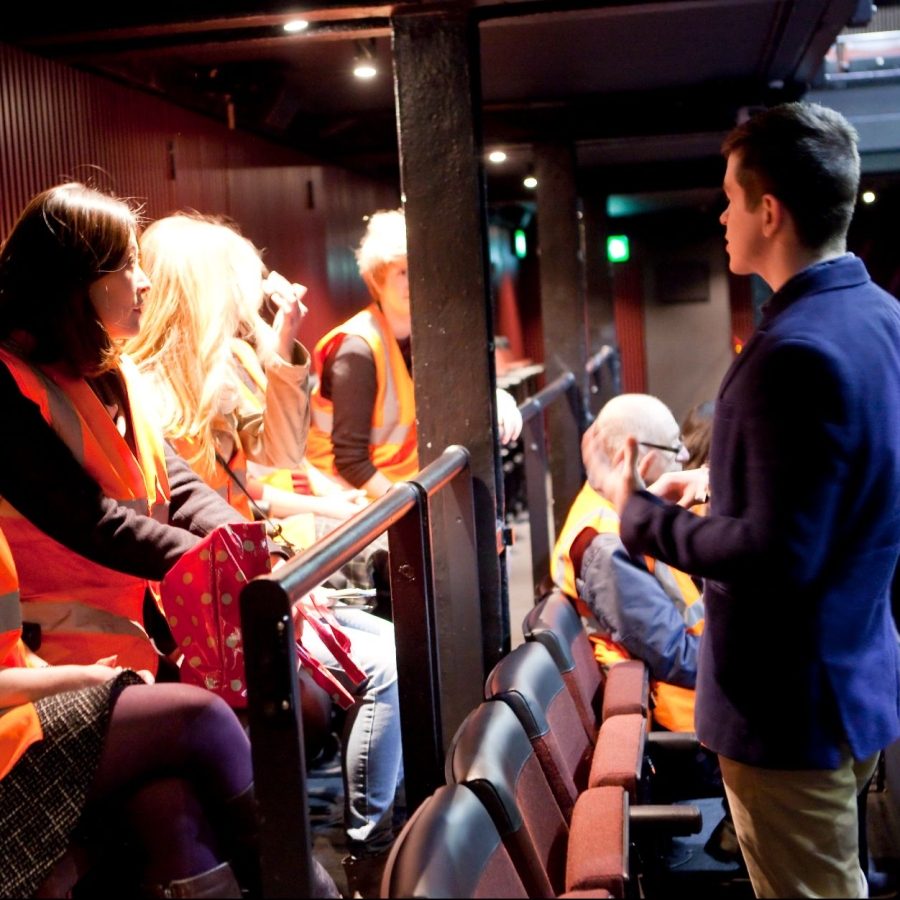 Backstage-Tour-Dorfman-Theatre A tour guide talking to a tour group in the Dorfman theatre. Tour participants wearing orange high-visibility vests.