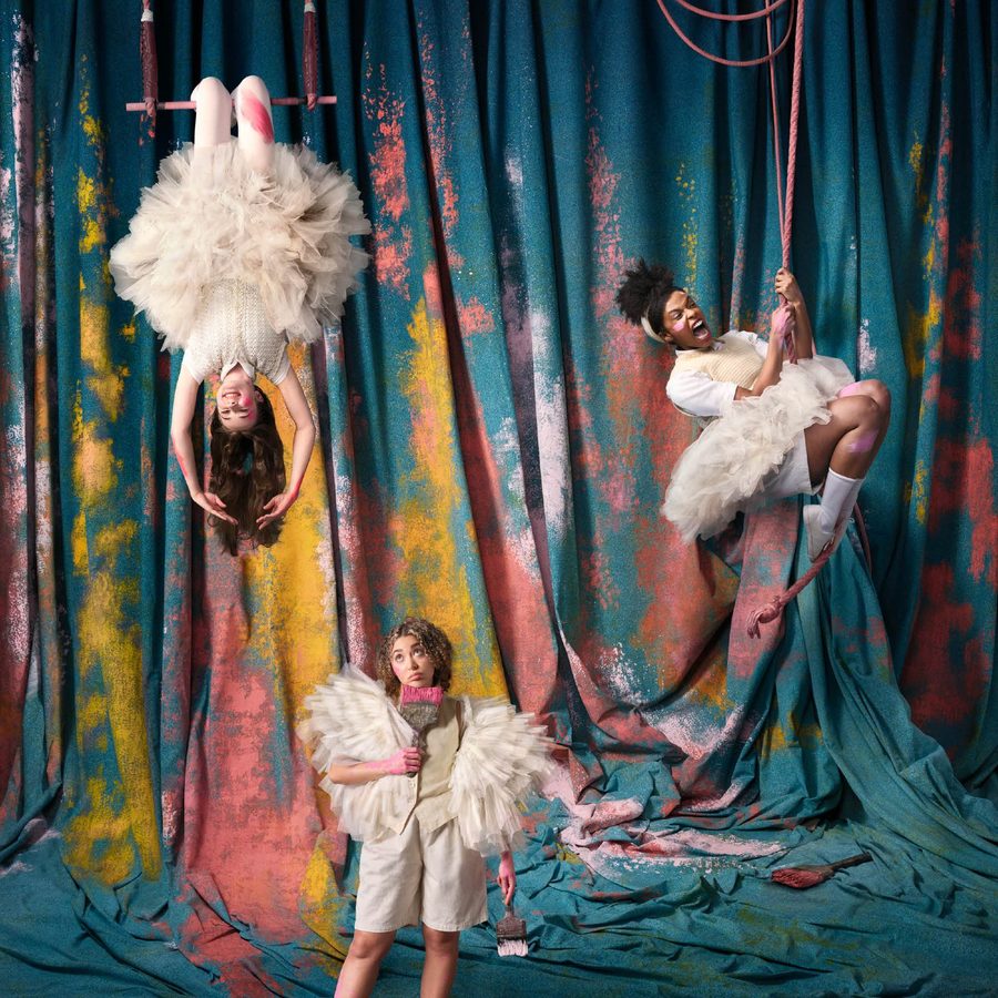 Ballet-Shoes-National-Theatre-2000x1000-v3 Three performers in whimsical costumes pose against a colorful, painted backdrop. One is upside down on a trapeze, another stands holding a prop, and the third hangs from aerial hoops. They wear fluffy dresses and expressive accessories.