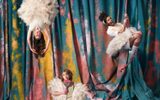 Ballet-Shoes-National-Theatre-1500x1500-v3 Three performers in whimsical costumes pose against a colorful, painted backdrop. One is upside down on a trapeze, another stands holding a prop, and the third hangs from aerial hoops. They wear fluffy dresses and expressive accessories.