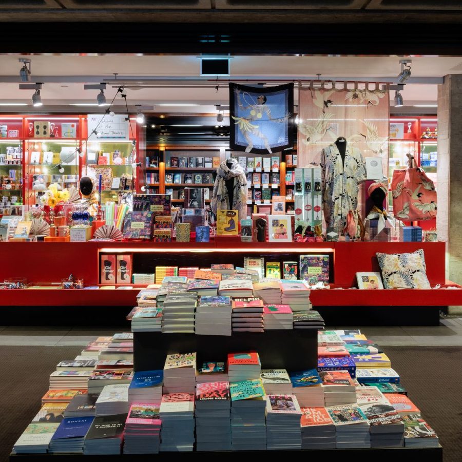 A brightly lit bookstore with numerous books on tables and shelves. The central display features colorful children's books and decorative items. Shelves on the sides are filled with books of various genres, and a large open space leads to the back of the store.
