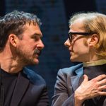Two men look at one another. One is wearing black glasses and a grey jacket and one is wearing a navy jacket.