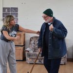 A man walks with a cane and wears a green beanie and denim shirt. A woman stands on his left and touches his arm while speaking to him.