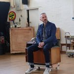 A man sits on a wooden box and wears a denim shirt.