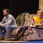 A man and a woman sit on a brown and black patterned sofa. The woman is wearing a yellow jumper and the man is wearing a grey jumper.