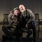 Two men in thick coats with smiles on their faces. The man on the left is sat on the floor and pointing forward. The man on the right is crouched down to put an arm around the first man's shoulders.