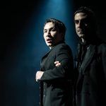 Two men in black coats under a spotlight. The man on the left is in the middle of the light, looking to the right with a shocked expression. He is being held by the arm by the man on the right who is more in shadow.