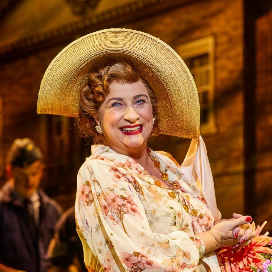 Caroline Quentin as Mrs Malaprop in Jack Absolute Flies Again Caroline Quentin as Mrs Malaprop, wearing a wide-brimmed hat and a big smile, holding a bunch of flowers, with airmen and a country house in the background.
