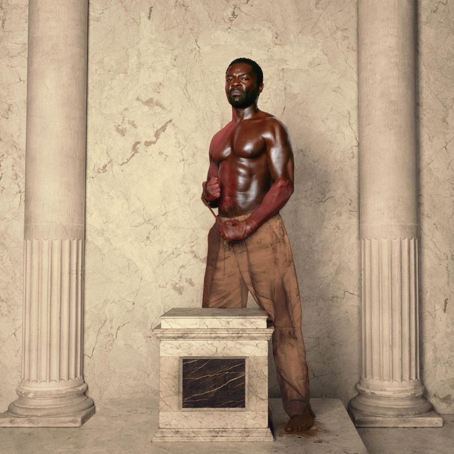 Coriolanus-David-Oyelowo-National-Theatre-v2-2000x1000 A shirtless man with a muscular build stands confidently between two stone columns. He has a defiant expression and has leather strapping wound around one hand and the wrist of his other arm. The background features a textured marble wall.