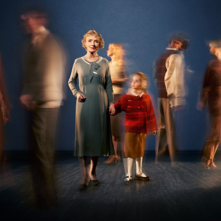 Dear-Octopus-Lindsay-Duncan-National-Theatre-v2-2000x1000 A woman in a brown long-sleeved dress stands, smiling, looking straight to camera. She is holding hands with a young girl who looks up at her. The blurred figures of five other people stand either side of them and the girl is also blurred.