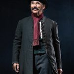 A man wearing an old fashioned coat a top hat and scarf, stands, smiling, holding a small suitcase.