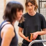 A woman with long brown hair wearing a black t-shirt and holding an orange purse. She is looking at another woman who is out of focus in the foreground, who has short brown hair and is wearing a blue tank top.