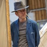 A man wearing a grey fedora, a denim jacket, and a grey shirt stands in front of a canvas tent. He has his hands behind his back and is looking to the side.