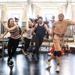 In a brightly lit room, a group of people are enjoying going through a dance routine. Behind them some people are standing on a raised platform.